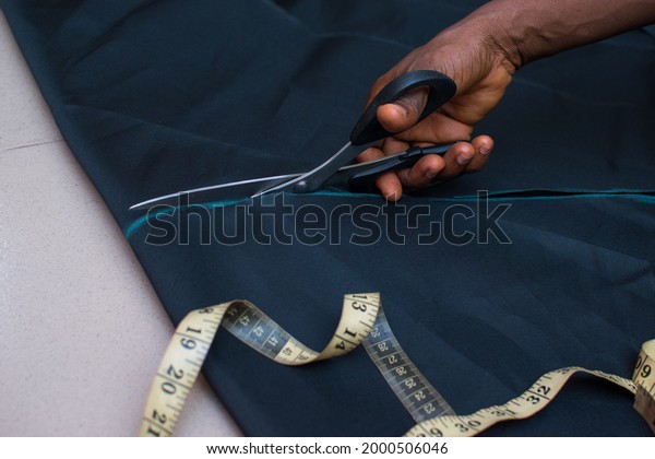 Hand of an African female tailor or fashion\
designer cutting a measured cloth with scissors to make desired\
dress in a tailoring\
workspace