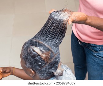 Hand of an African stylist, holding, displaying and applying relaxer cream to the natural long hair of a woman or female customer at a beauty salon in Nigeria - Powered by Shutterstock