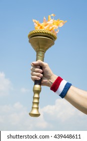 Hand Of An American Athlete Wearing USA Red, White, And Blue Colors Sweatband Holding Stylized Flame Against Tropical Blue Sky