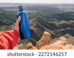 Hand of anonymous hiker holding a blue metal water bottle on a rocky cliff.
