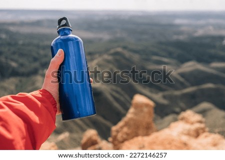 Hand of anonymous hiker holding a blue metal water bottle on a rocky cliff.