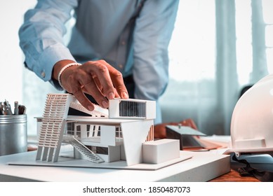 Hand of architect-designer holding the part of the model of modern box house while thinking about concept of building and construction project in the future. Focusing on his hand holding model. 