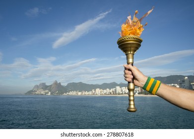 Hand Of An Athlete Wearing Brazil Colors Sweatband Holding Sport Torch Against Rio De Janeiro Brazil Skyline With Two Brothers Mountain