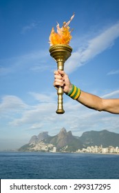 Hand Of An Athlete Wearing Brazil Colors Sweatband Holding Sport Torch Against Rio De Janeiro Brazil Skyline With Two Brothers Mountain