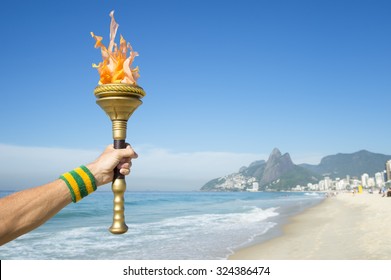 Hand Of An Athlete Wearing Brazil Colors Sweatband Holding Sport Torch On Ipanema Beach With Two Brothers Mountain On The Skyline Of Rio De Janeiro, Brazil