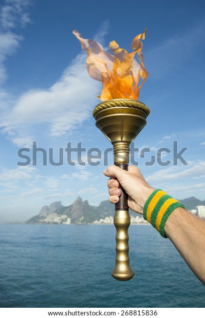 Hand of an athlete wearing Brazil colors\
sweatband holding sport torch against Rio de Janeiro Brazil skyline\
with Two Brothers Mountain
