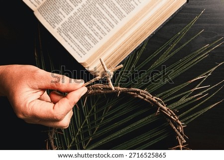 Hand attaching small palm leaf cross to crown of thorns above open Bible with palm leaves, symbolizing Christian faith, Palm Sunday ritual, sacrifice and Easter reflection for worship, prayer concepts