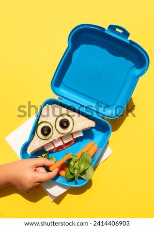 Hand of baby girl picking up baby carrot from lunch box with funny looking sandwich with anthropomorphic face