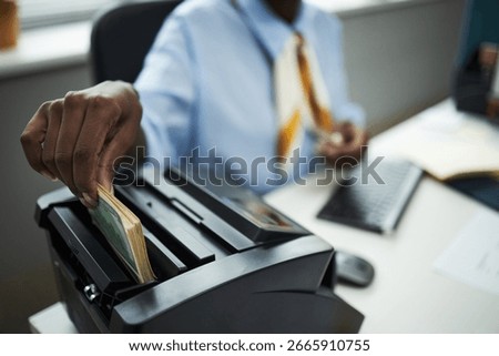 Hand of bank worker processing stack of cash using bill counting machine in office, hand inserting banknotes while sitting at desk with computer keyboard and documents visible