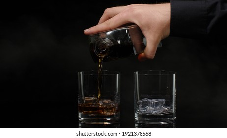 Hand of barman pouring golden whiskey, cognac or brandy from bottle into glasses with ice cubes on black background. Bartender pouring glass of alcohol drink bourbon, rum. Copy-space