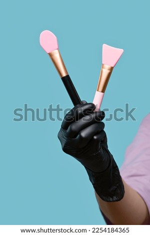 Hand of Beautician holds set of spatulas for applying cosmetic masks. Empty space. Cosmetologist in black latex glove on blue background in studio