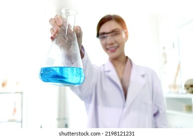 Hand Of Beautiful Asian Female Scientist Wear Lab Coats And Safety Glasses, Happy Smiling Researcher Woman Standing And Showing Blue Chemical Flask In Laboratory, Woman Work  In Science Field.