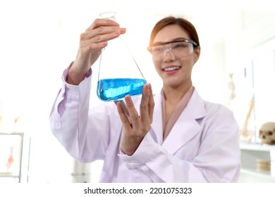 Hand Of Beautiful Asian Female Scientist Wear Lab Coats And Safety Glasses, Happy Smiling Researcher Woman Standing And Showing Blue Chemical Flask In Laboratory, Woman Work  In Science Field.