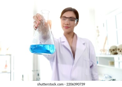 Hand Of Beautiful Asian Female Scientist Wear Lab Coats And Safety Glasses, Happy Smiling Researcher Woman Standing And Showing Blue Chemical Flask In Laboratory, Woman Work  In Science Field.