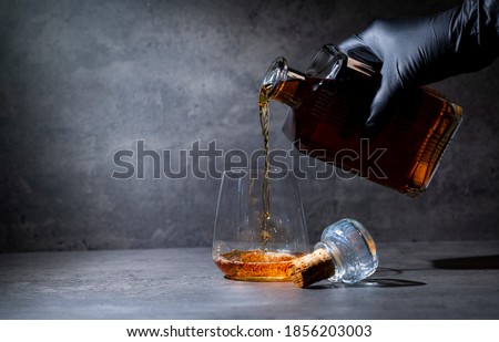 hand in a black glove pours whiskey from a square bottle into a glass on a dark gray cement background