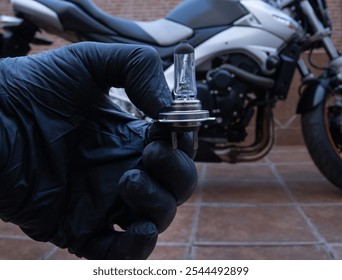 A hand with black latex gloves, holding a halogen motorcycle bulb. A motorcycle in the background. Maintenance concept. - Powered by Shutterstock