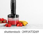 Hand blender with mixture of ingredients and fresh fruits on white marble table, space for text