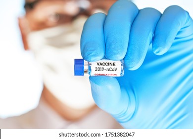 Hand In Blue Medical Gloves Holding Small Vial With Label (own Design, Not Real Product) 2019-nCoV Vaccine, Blurred Face In Cotton Mask And Goggles Background. Coronavirus Covid 19 Cure Concept