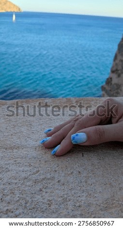 Hand with blue patterned manicure resting on a stone ledge overlooking clear turquoise sea, with a blurred coastline and sailboat in the background under a bright sky.