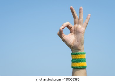 Hand of Brazilian athlete making OK sign with finger and thumb against blue sky wearing a Brazil colours yellow and green sport wristband
