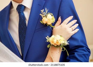 
The hand of the bride with a boutonniere rests on the forearm of the groom in a blue suit with a boutonniere and tie