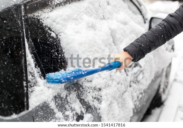 hand with brush removing snow from car in winter\
day. woman cleaning snow from car. woman is sweeping off snow from\
her car after snowfall