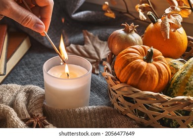 Hand With Burning Match Lighting A Candle On The Windowsill With Cozy Autumn Still Life With Pumpkins, Knitted Woolen Sweater And Books. Autumn Home Decor. Cozy Fall Mood. Thanksgiving. Halloween.