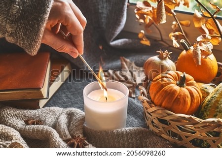 Hand with burning match lighting a candle on the windowsill with cozy autumn still life with pumpkins, knitted woolen sweater and books. Autumn home decor. Cozy fall mood. Thanksgiving. Halloween.