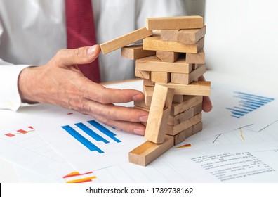 Hand Of Businessman Catching Wooden Blocks Of Wooden Tower Jenga Falling Down On Table Full Of Graphs Diagrams Charts. Crisis And Analyzing Concept