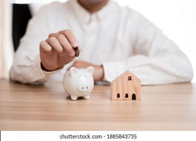  Hand Of Businessman Putting Coin In Little Piggy Bank And Small Wooden Toy House Placed On The Table, The Idea Of ​​ Saving Money To Buy A House For A Better Quality Of Life For Family, Saving Energe