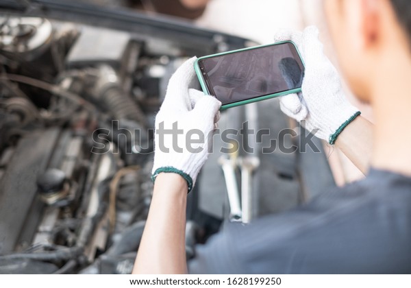 Hand of car\
mechanic or technician holding smartphone taking photography of car\
engine open hood. Car service\
concept.