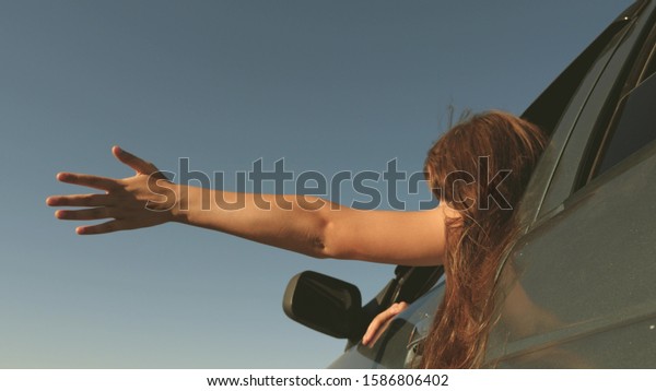 hand from car window catches the wind. Girl with\
long hair is sitting in front seat of car, stretching her arm out\
window and catching glare sun. happy female traveler rides car\
along country road.