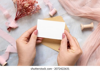 Hand With A Card And Envelope Over Marble With Dried Pink Flowers And Silk Ribbons Top View. Flat Lay With Horizontal Blank Card. Romantic Thank You Or Rsvp Card Mockup, Copy Space