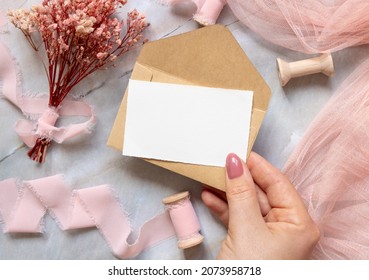Hand with a card and envelope over marble with dried pink flowers and silk ribbons top view. Flat lay with horizontal blank card. Romantic Thank you or rsvp card mockup, copy space