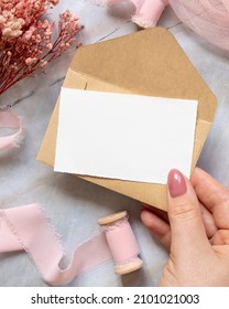 Hand with a card and envelope over marble with dried pink flowers and silk ribbons top view. Flat lay with horizontal blank card. Romantic Thank you or rsvp card mockup, copy space