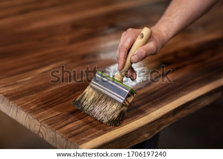Hand of a carpenter designer oiling an authentic self made table with a big brush in a workshop environment. Finishing beautiful teak wood slabs with protective oil