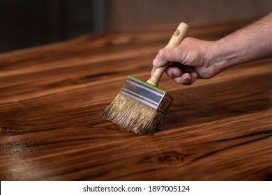 Hand Of A Carpenter Oiling An Authentic Self Made Table With A Big Brush In A Workshop Environment. Finishing Beautiful Teak Wood Slabs With Protective Oil