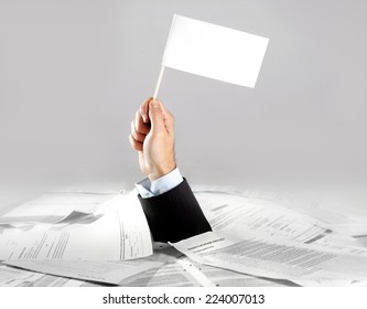 Hand Of Caucasian Businessman Emerging From Office Desk Loaded Of Paperwork , Invoices And A Lot Of Papers And Documents Holding White Flag  Overworked In Giving Up Concept