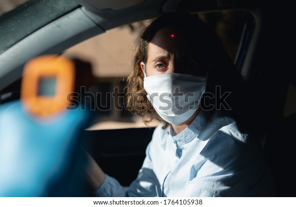 Hand of a Caucasian\
man taking temperature of a Caucasian woman sitting in her car and\
wearing a face mask