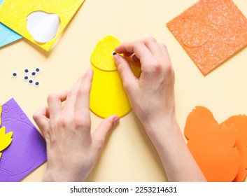 The hand of a caucasian teenage girl glues a decorative eye on a felt yellow chick, sitting at a table with a set of craft objects on a pale yellow background, close-up flat lay. 