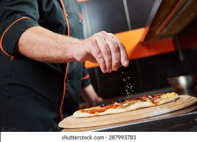 hand of chef baker in uniform adding spice into pizza after pizza preparation at restaurant kitchen