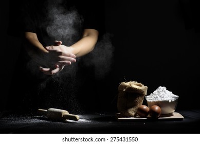 hand of chef clap a flour with wooden rolling pin and ingredients on black background