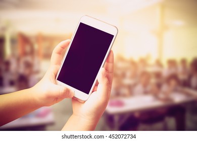 Hand Of Child Holding Mobile Phone With Blur Student Learning In Classroom Background