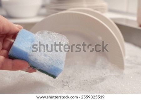 Hand with cleaning sponge with soapy foam. Kitchen sink filled with water and detergent. Washing dishes.