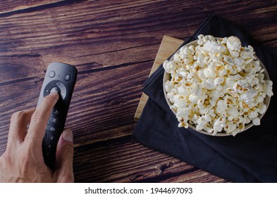 a hand clicking a TV remote control with a bowl of popcorn placing on the side on a table cloth on dark brown wood table - Powered by Shutterstock