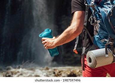 Hand close up with drinking water bottle. Man with backpack dressed in active trekking clothes touristic staying near mountain river waterfall and enjoying Nature. Traveling, trekking concept image