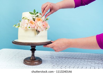 Hand Close-up Decorates Working Space Baker Confectioner Pastry Chef Appetizing Creamy White Two-tiered Wedding Cake Decorated With Fresh Flowers On A Table Studio On A Blue Background