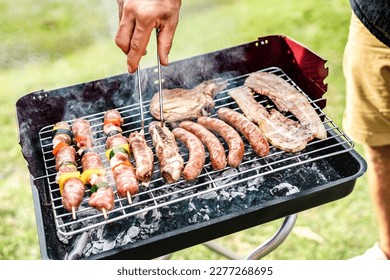 Hand closeup grilling meat at home bar-b-q session - Skewers and burger sausages cooked on ember at bbq pinic party - Pic nic life style concept with lunch out side on spring summer time - Warm filter - Powered by Shutterstock