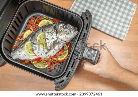 HAND COOKING GILTHEAD SEA BREAM OR DORADO WHOLE FISH WITH GARNISH IN AIR FRYER IN THE KITCHEN. KETO DIET FOOD RECIPES. TOP VIEW.