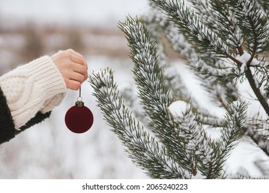 Hand decorating pine tree branches with modern red bauble. Decorating christmas tree in snow outdoors. Preparation for winter holidays in countryside. Merry Christmas! Space for text - Shutterstock ID 2065240223
